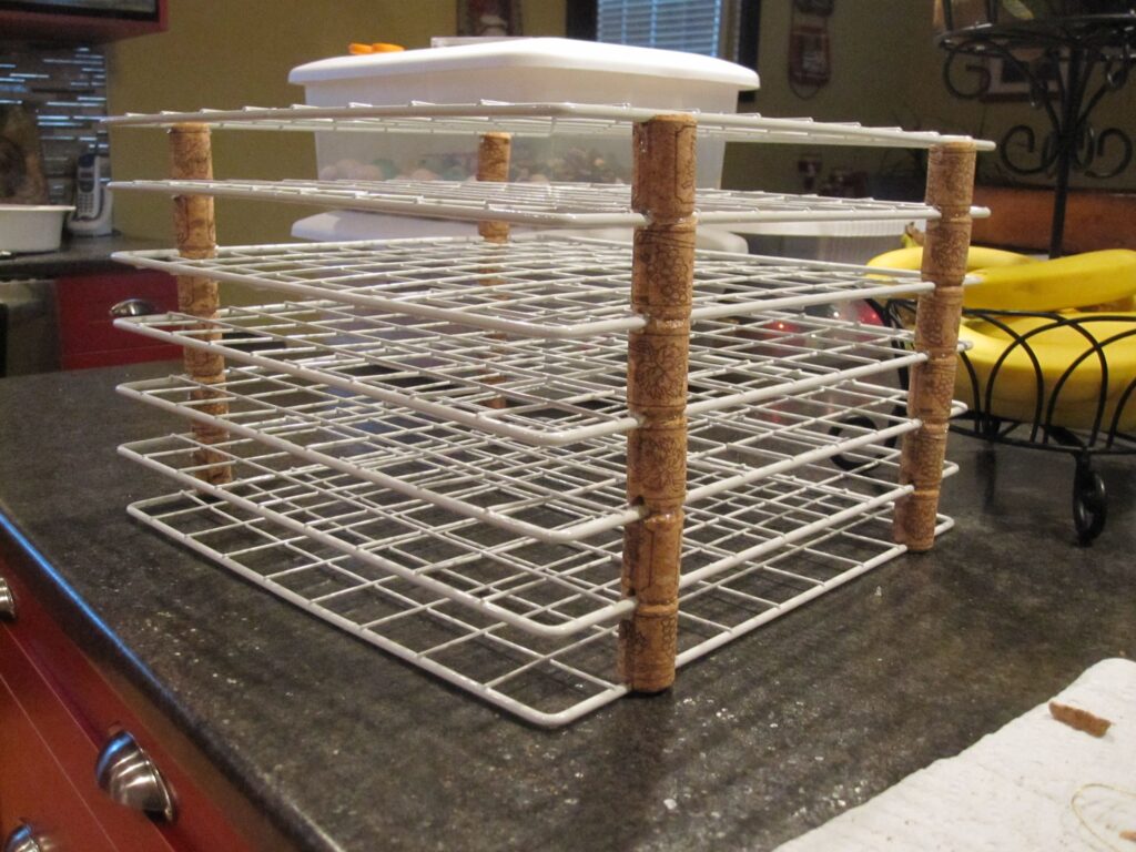A multi-level homemade drying rack constructed from wire shelves and cork spacers. It stands on a kitchen countertop, with items like bananas, a white-lidded container, and kitchen utensils visible in the background. The setup appears DIY and functional, likely used for drying art. 