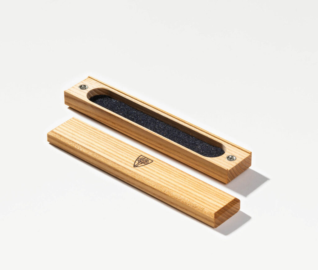 A rectangular wooden dice carrier with a detachable lid, crafted from light-colored wood. Inside, black foam padding snugly holds polyhedral dice. The lid, resting beside the case, features an engraved triangular emblem. The carrier is secured with metal screws at each end, giving it a clean, functional, and portable design for tabletop gaming.