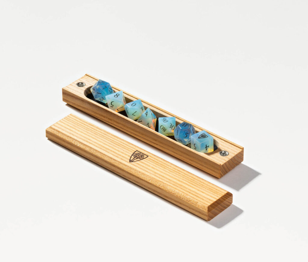 An open wooden dice case displaying a full set of polyhedral dice in varying shades of blue. The dice, commonly used in tabletop games like Dungeons & Dragons, are neatly arranged inside the case. The detached lid lies beside it, featuring a sleek logo or emblem.