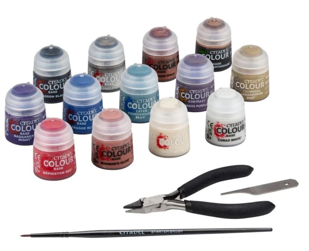 A hobby miniature painting starter set featuring 13 Citadel Colour paint pots in various shades, a black-handled detail brush labeled "Starter Brush," a pair of black-handled clippers, and a metal hobby knife blade. The paint selection includes base, layer, contrast, shade, and technical types, commonly used for Warhammer miniatures and other tabletop models.
